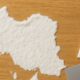 This image shows a white powdery substance shaped like the map of Iran on a wooden surface. To the right of the shape are a white cylindrical object resembling a straw and a metallic rectangular card, suggestive of items often associated with drug use. Both this description and this image were generated using artificial intelligence.