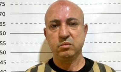 Wassim Badi al-Assad, a bald man wearing a striped prison uniform stands against a height chart with measurement lines in the background, facing the camera with a neutral expression. Description generated with AI.