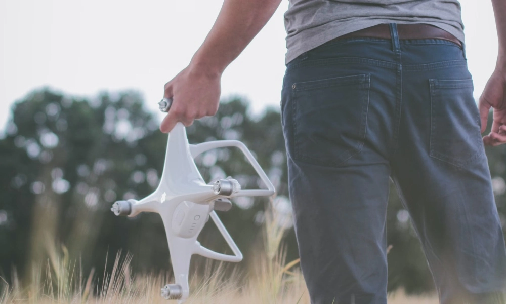A person stands in a grassy field holding a white quadcopter drone by one of its arms, with their back turned to the camera. The image captures a relaxed, outdoor moment likely just before or after a drone flight. (description generated with AI)