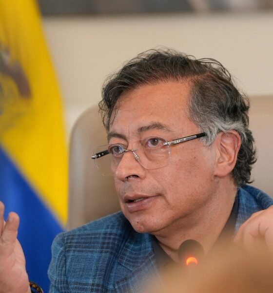 Gustavo Petro in a blue plaid jacket and glasses speaks into a microphone with a serious expression and hand gesture, seated in front of a blurred Colombian flag. The setting suggests an official or political context, likely a press conference or government meeting.