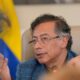 Gustavo Petro in a blue plaid jacket and glasses speaks into a microphone with a serious expression and hand gesture, seated in front of a blurred Colombian flag. The setting suggests an official or political context, likely a press conference or government meeting.
