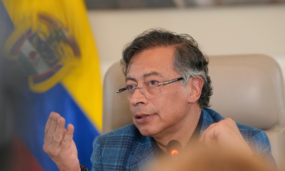 Gustavo Petro in a blue plaid jacket and glasses speaks into a microphone with a serious expression and hand gesture, seated in front of a blurred Colombian flag. The setting suggests an official or political context, likely a press conference or government meeting.